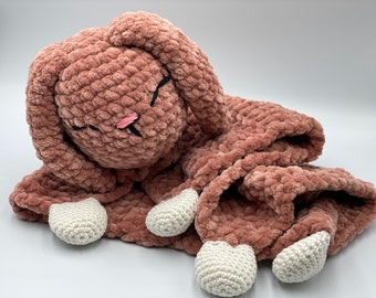 May include: A soft, plush pink crocheted bunny with white crocheted feet. The bunny is lying on its side with its head resting on its paws.