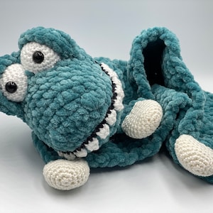 May include: A teal blue crocheted alligator scarf with white and black accents. The scarf has large white eyes and a white crocheted pom-pom on each end.