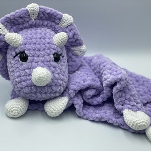 May include: A purple crocheted triceratops soft toy with white accents. The triceratops is attached to a purple crocheted blanket with white accents.