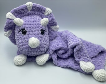 May include: A purple crocheted triceratops soft toy with white accents. The triceratops is attached to a purple crocheted blanket with white accents.