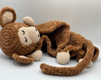 May include: A brown crocheted monkey toy with a beige face and closed eyes. The monkey is lying down with its tail curled around its body. The toy is made with a soft, plush yarn.
