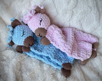 Puede incluir: Dos mantas de seguridad hechas a mano con ganchillo, una azul claro y otra rosa, cada una con una cabeza de caballo y detalles marrones. Las mantas son suaves y afelpadas, perfectas para un bebé o niño pequeño. El fondo es una superficie texturizada de color crema.