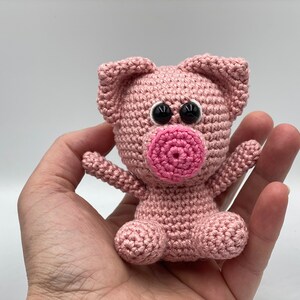 May include: A pink crocheted pig toy with black eyes and a pink snout. The pig is sitting in a hand.