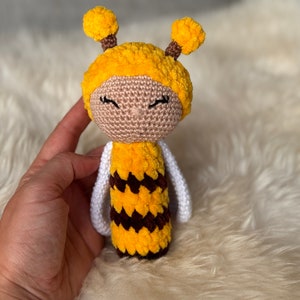 May include: A handmade crochet doll in the shape of a bee. The doll has a yellow fuzzy head with two small antennae, a tan face, and closed eyes. The body is yellow with brown stripes, and it has white wings.