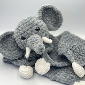 Puede incluir: Un juguete de peluche de elefante de crochet gris con detalles de crochet blanco. El elefante está unido a una manta de crochet gris.