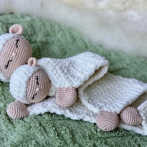 Puede incluir: Dos peluches de oveja blancas de crochet con caras rosas y ojos cerrados. La oveja más grande está tumbada de espaldas con las patas en el aire, y la oveja más pequeña está tumbada de lado con la cabeza apoyada en el cuerpo de la oveja más grande. Los peluches están sobre una superficie verde peluda.