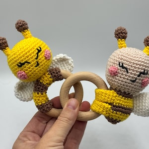 May include: A handmade crochet baby rattle featuring two smiling bee figures in yellow, brown, and white, with a wooden ring. The bees have pink cheeks and closed eyes. A charming and safe toy for infants.