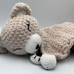 Pode incluir: Um urso de pelúcia de crochê bege com detalhes brancos, deitado de lado com a cabeça apoiada na pata. O urso tem os olhos fechados e um nariz de crochê branco.