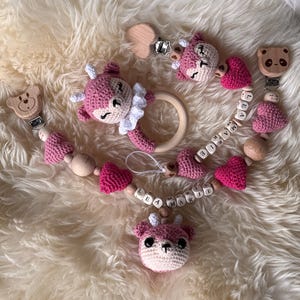 Op de afbeelding: Een verzameling handgemaakte babyaccessoires, waaronder een fopspeenclip, een houten ring en een fopspeenhouder. De items hebben roze gehaakte herten- en hartvormen, houten kralen en beer-vormige clips. De naam "MARIE" staat op de fopspeenclip.