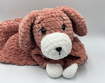 May include: A crocheted plush dog toy in pink and white. The dog is lying down with its head resting on its paws. The toy has a white face with black eyes and a black nose.