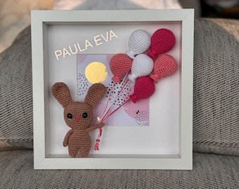 May include: A brown crocheted bunny holding a bunch of pink and white crocheted balloons. The balloons are attached to a white frame with the text "PAULA EVA" on it.