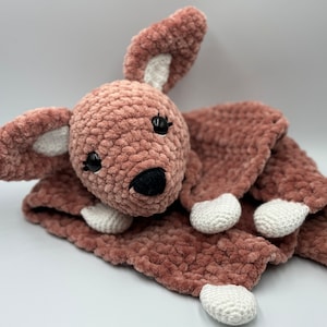 Puede incluir: Un juguete de peluche de canguro de crochet rosa y blanco con ojos negros y nariz negra. El canguro está acostado de lado con las piernas extendidas.