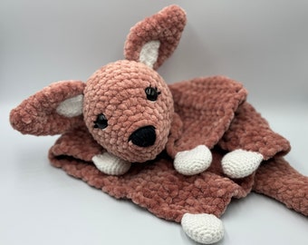 May include: A crocheted pink and white kangaroo plush toy with black eyes and a black nose. The kangaroo is lying on its side with its legs outstretched.
