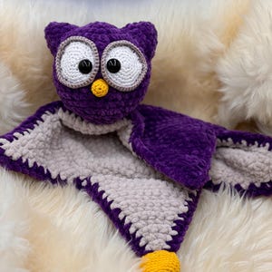 Könnte beinhalten: Lilafarbener gehäkelter Eulen-Schmusetuch mit grauem gehäkeltem Rücken und gelben Füssen.