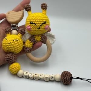 May include: A handmade baby pacifier clip featuring a yellow crocheted bee design with brown accents and a wooden ring. The clip includes the name "SOPHIA" spelled out with wooden letter beads. The bee has pink cheeks and white wings.