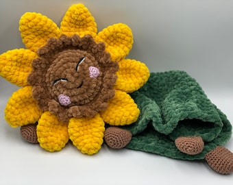 May include: A crocheted sunflower with a brown face and pink cheeks, paired with a green crocheted blanket. The sunflower has a happy expression.