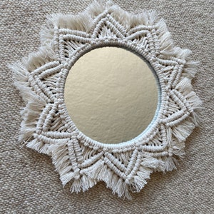 May include: A white macrame mirror with a starburst design and fringe trim. The mirror is round and has a white background.