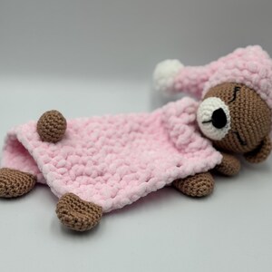 May include: A handmade crochet baby comforter featuring a brown bear head wearing a pink sleep cap. The bear is lying on a pink blanket with brown paws. The comforter is designed for babies and young children.