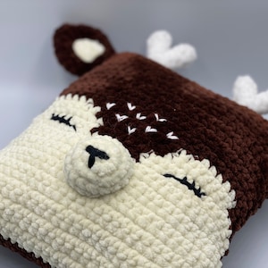May include: A brown and white crocheted deer cushion with a white fluffy texture. The deer has a brown nose and black eyes.