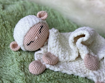 May include: A crocheted white lamb with a pink face and closed eyes is lying on a green fuzzy blanket. The lamb is holding a white crocheted blanket.