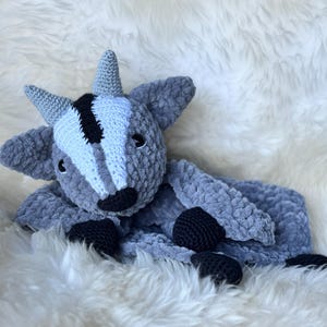 May include: A gray and white crocheted goat plush toy with black eyes and nose. The toy is lying on a white fluffy surface.