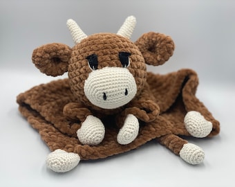 May include: A brown crocheted cow plush toy with white accents. The cow is attached to a brown crocheted blanket.