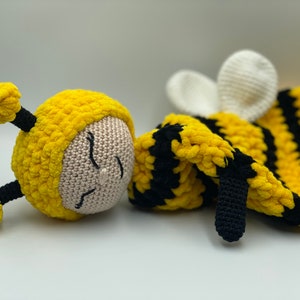 Puede incluir: Un juguete de peluche de abeja tejido a crochet con rayas amarillas y negras. La abeja tiene una cara blanca con ojos bordados y una boca cerrada.