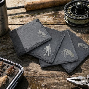 May include: Four square slate coasters with engraved fishing fly designs. The coasters are arranged on a wooden surface, alongside fishing gear, including a reel, a fly box, and a stainless steel tumbler with the text "DRINK COFFEE CATCH FISH."