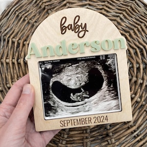May include: A wooden frame with a light brown background and a white border. The frame has a cutout in the shape of an arch. The text "baby Anderson" is written in green letters on the top of the frame. The text "SEPTEMBER 2024" is written in black letters on the bottom of the frame. A black and white ultrasound image is displayed in the center of the frame.