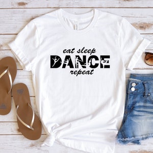 May include: White t-shirt with black text that reads "eat sleep dance repeat" with a silhouette of a dancer in each letter of the word "dance". The shirt is laid out on a wooden surface with a pair of brown flip flops and blue denim shorts.