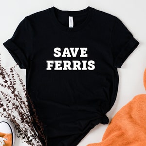 May include: Black t-shirt with white text that reads "Save Ferris". The shirt is laid flat on a white surface with a sprig of dried flowers and a pair of orange trainers.