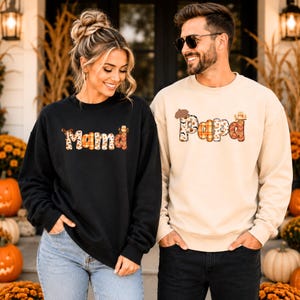 May include: A black sweatshirt with the word Mama in a fall-themed patterned font and a beige sweatshirt with the word Papa in a matching fall-themed patterned font, both featuring autumn motifs like leaves, pumpkins, and small woodland animals.