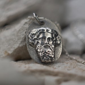 Op de afbeelding: Een zilveren ketting met een hanger met een gedetailleerd, gegraveerd portret van een man met een baard. De hanger is ovaalvormig en hangt aan een zilveren ketting.
