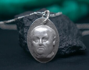 May include: A silver pendant necklace featuring a detailed, three-dimensional portrait of a person's face.