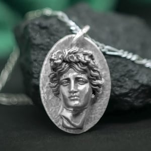 Puede incluir: Colgante ovalado plateado con un busto clásico con una corona de laurel. El colgante cuelga de una delicada cadena de plata. El fondo incluye una piedra oscura y un tejido verde borroso.