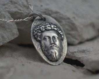 May include: A silver pendant necklace featuring a detailed, embossed image of a man's face with a beard and curly hair. The pendant is oval-shaped and hangs from a silver chain.