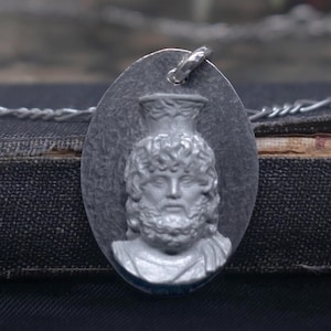 Puede incluir: Colgante ovalado de plata con un busto de un hombre con barba y pelo rizado, que lleva un casco. El colgante está suspendido de una cadena de plata. El fondo es una superficie oscura y texturizada.