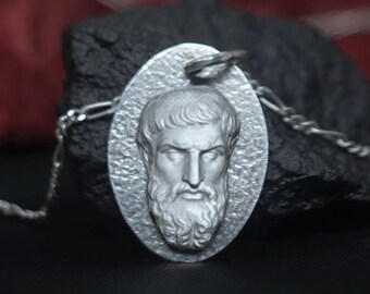 May include: Silver-toned pendant necklace featuring an oval pendant with a detailed relief of a bearded man's face. The pendant is attached to a delicate chain. The background is a dark, textured surface.