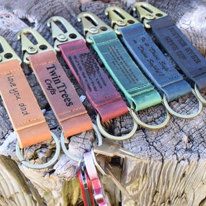 May include: A set of six leather keychains with different engraved messages. The keychains are brown, red, green, blue, and black. The keychains are made of leather and have a metal clasp. The keychains are all on a wooden surface.