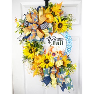 Könnte beinhalten: Ein Herbstkranz mit Sonnenblumen, Kürbissen und einem blau-weiß gemusterten Schild mit der Aufschrift "Willkommen Herbst". Der Kranz ist mit einer gelben und blauen Schleife verziert.
