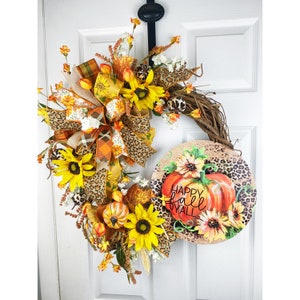Könnte beinhalten: Ein Herbstkranz mit einer braunen Weinrebenbasis, verziert mit Sonnenblumen, Kunstblättern und einer Schleife im Leopardenmuster. Ein rundes Schild mit dem Text "Happy Fall Y'all" und einem bemalten Kürbis ist am Kranz befestigt.
