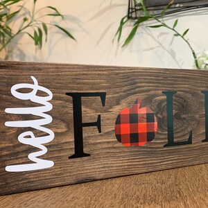 Puede incluir: Un letrero de madera con el texto "hello fall" en letras blancas en cursiva, una "F" y una "LL" negras, y una manzana de cuadros rojos y negros.