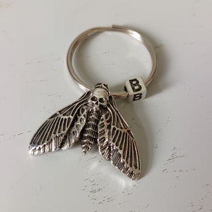 May include: A silver key chain with a detailed skull moth charm and a silver charm with the letter 'B' on it.
