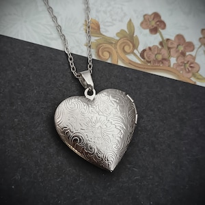May include: Silver heart-shaped locket necklace with an intricate floral design. The locket hangs from a delicate chain. The locket is closed, and the surface is reflective. The background is a mix of black and patterned paper.