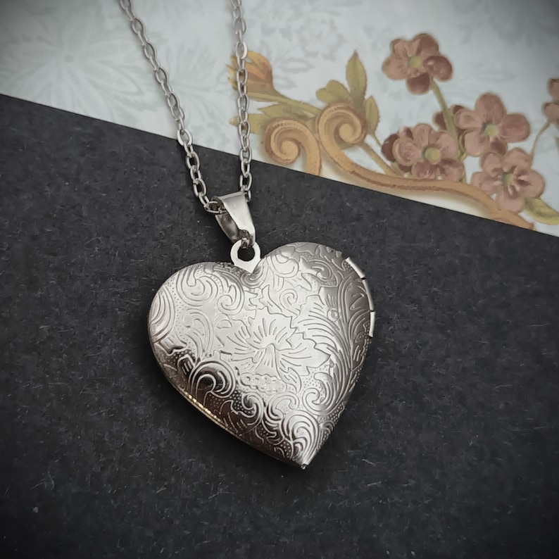 May include: Silver heart-shaped locket necklace with an intricate floral design. The locket hangs from a delicate chain. The locket is closed, and the surface is reflective. The background is a mix of black and patterned paper.