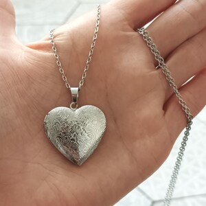 May include: Silver heart-shaped locket necklace with an intricate floral design. The locket hangs from a delicate silver chain. The heart pendant is approximately 1.5 inches wide and 1.5 inches tall.