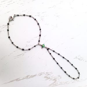 May include: A black beaded bracelet with a silver clasp and a green and silver charm. The bracelet is on a white wooden surface.