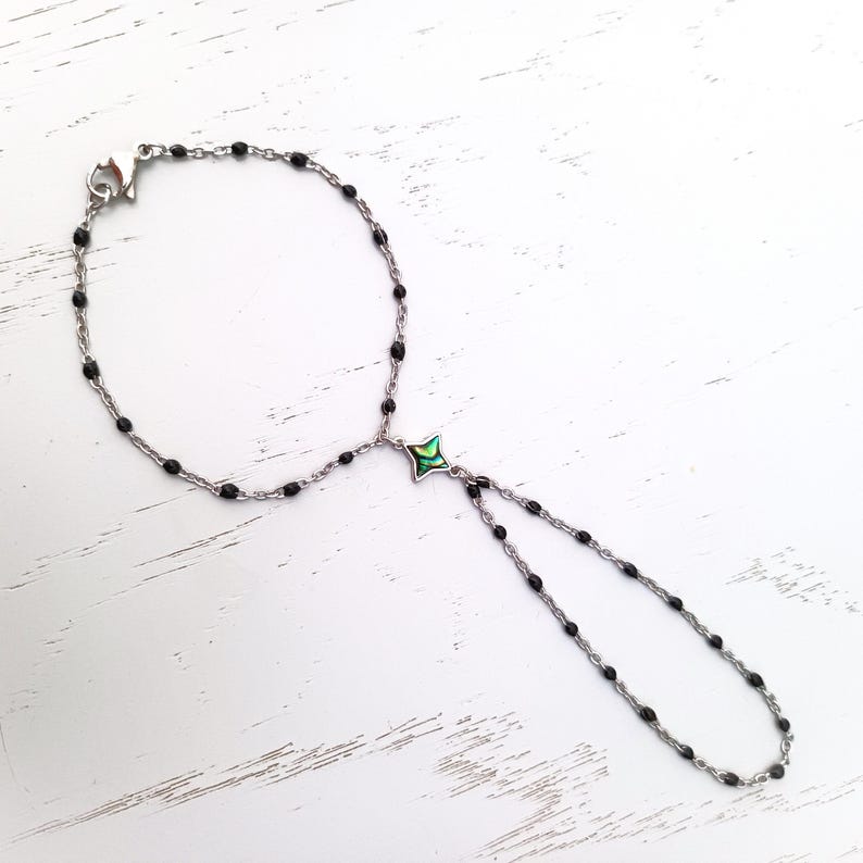 May include: A black beaded bracelet with a silver clasp and a green and silver charm. The bracelet is on a white wooden surface.