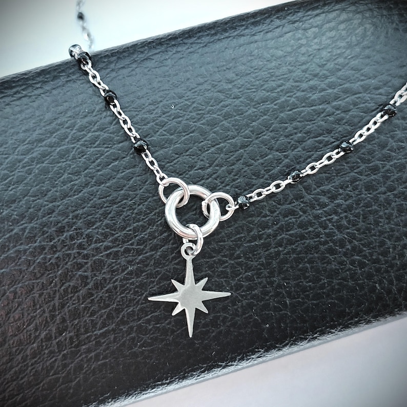Puede incluir: Un collar de cadena de plata con cuentas negras y un colgante en forma de estrella. La cadena tiene un cierre circular.