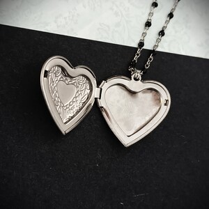 May include: Open silver heart-shaped locket necklace with a black beaded chain. The locket is open, revealing a smaller heart shape inside and a blank space for a photo. The locket rests on a black and white patterned surface.